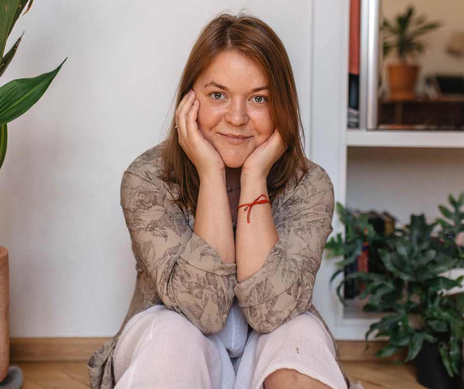 A woman looks confidently into the camera. She sits on a step with plants either side of her.