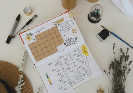 Image of someone creative journalling, planning their month ahead with teaxt and images in a plain notebook. Around the bok are pens, candles and lavender stems.