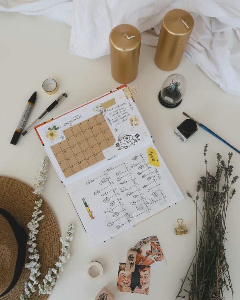 Image of someone creative journalling, planning their month ahead with teaxt and images in a plain notebook. Around the bok are pens, candles and lavender stems.