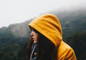 A woman stands in the countryside on a misty day, her yellow hood is up and she looks into the distance.